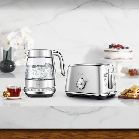 Silver kettle and toaster on a kitchen counter with a cake and tea set in the background.