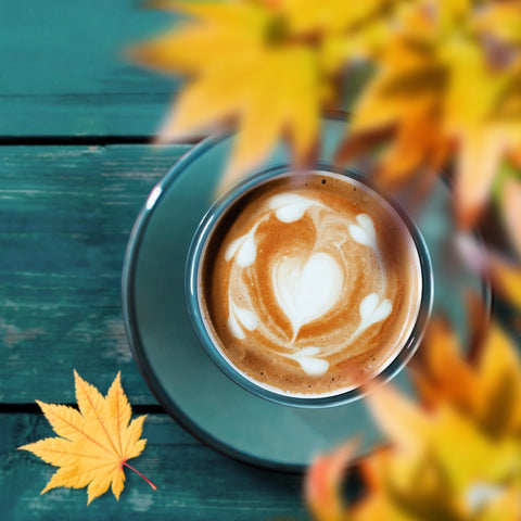 Cappuccino with latte art on a saucer, surrounded by autumn leaves and flowers on a wooden surface.
