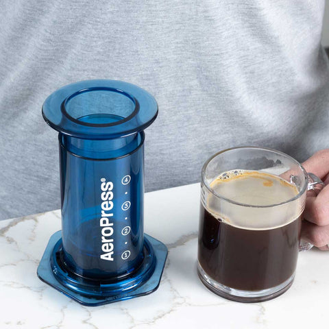 Blue AeroPress coffee maker next to a glass of coffee on a marble surface