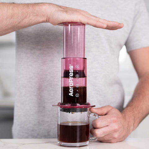 Person using an Aeropress coffee maker with a glass of coffee on a table.