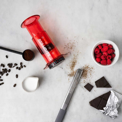 Red aeropress maker on a marble surface with coffee beans, raspberries, and chocolate.
