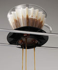 Coffee dripper with Kalita Wave 185 Glass Dripper and coffee dripping into a black container on a gray background.