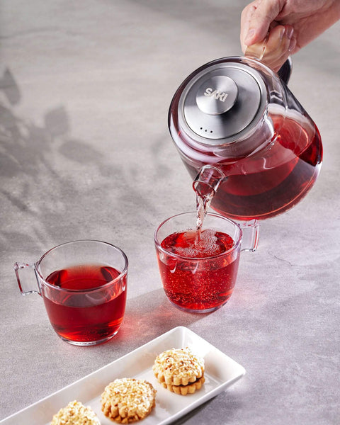 Person pouring red tea from a glass teapot into a cup on a gray surface with cookies.