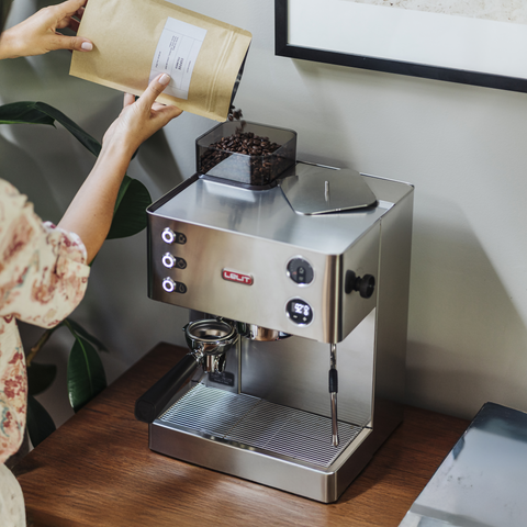 Person loading coffee beans into a Lelit Kate espresso machine on a wooden surface.