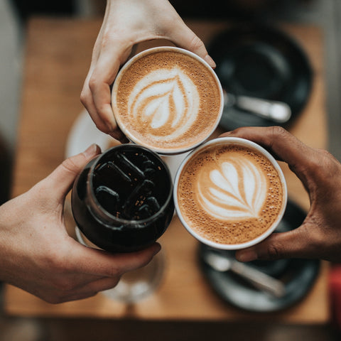 Three hands holding cups of coffee with latte art on a wooden table.