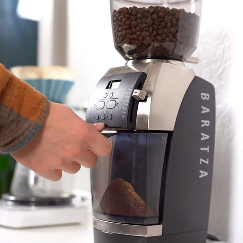 Baratza Vario™ W+ coffee grinder with a hand adjusting the setting, surrounded by blurred kitchen items.
