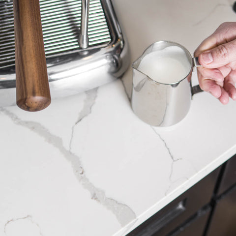 Person pouring milk from a metal pitcher into a Ascaso Dream PID Automatic Home Espresso Machine - Love Red  on a marble countertop.