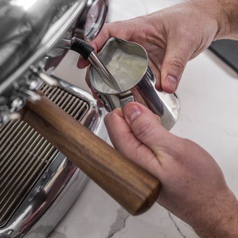 Person using a milk frother to froth milk in front of a coffee machine.