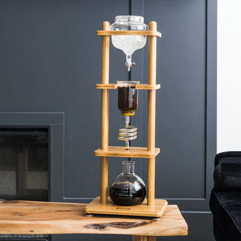 Bamboo coffee brewing apparatus with glass containers on a wooden stand against a gray wall.