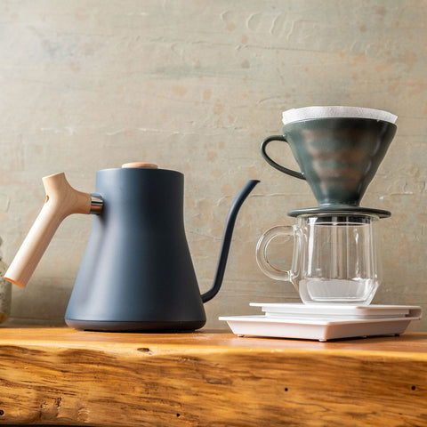 Coffee-making setup with a kettle, Hario V60 Ceramic Coffee Dripper 02 - Juniper, and glass mug on a wooden surface.