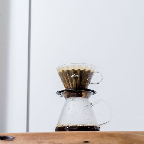 Coffee brewing device with Kalita Wave 185 Glass Dripper  on a wooden surface and light gray background