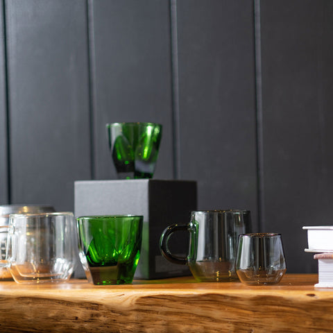 Set of green and clear glass mugs on a wooden surface with a dark paneled wall background.