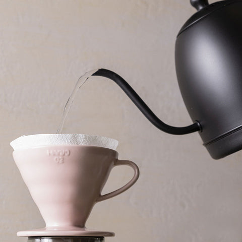Black kettle pouring water into a Hario V60 Ceramic Dripper in Pink over a white cup on a beige background