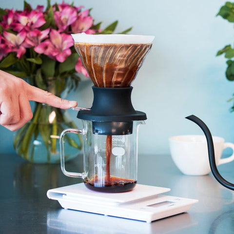 Coffee brewing setup with a hand pointing at a glass coffee maker on a kitchen counter.