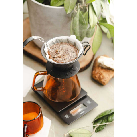 Coffee brewing setup with a Hario V60 Metal Coffee Dripper 02 - Black on a digital scale, surrounded by a plant and toast.