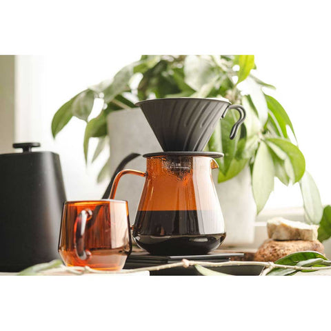 Coffee brewing setup with a glass carafe and Hario V60 Metal Coffee Dripper 02 - Black on a surface with plants in the background.