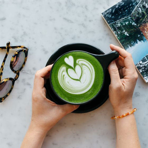 Person holding a cup of green matcha latte with a heart design on a marble surface.