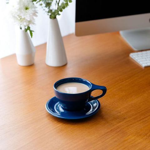 Blue cup of coffee on a wooden desk with a computer monitor and flowers in the background