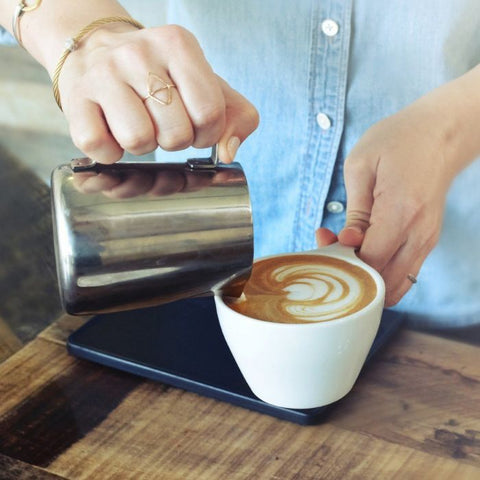 Person pouring milk into a cup of coffee with a latte art design on a wooden table.