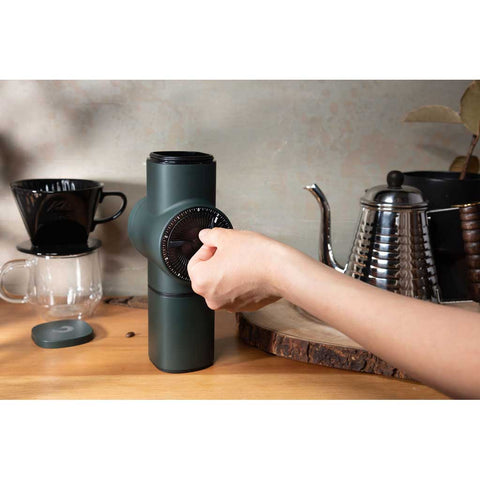 Person using a green coffee grinder on a kitchen counter with coffee-making equipment.