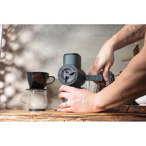 Person using a manual coffee grinder with a coffee filter and glass container on a wooden surface.