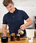Man pouring coffee into a glass with a Toddy Cold Brew System - Original Cold Brew coffee maker and grinder in the background.