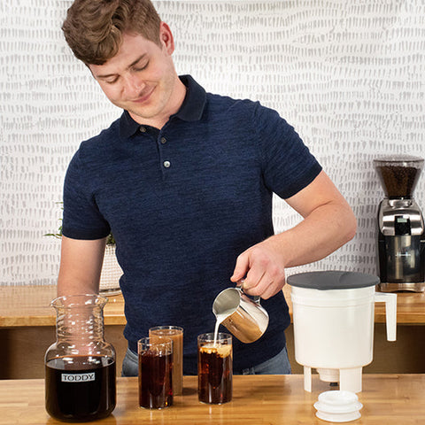 Man pouring coffee into a glass with a Toddy Cold Brew System - Original Cold Brew coffee maker and grinder in the background.