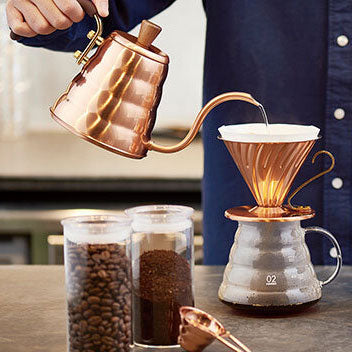 Person pouring coffee from a copper kettle into a ceramic dripper on a wooden table.