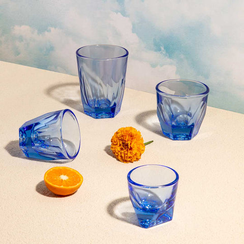 Blue glass tumblers and an orange on a sandy surface with a blue sky background