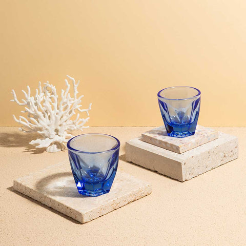Two blue glass cups on stone coasters with a piece of white coral against a beige background