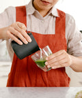 Person pouring green liquid from a black container into a glass, wearing a red apron.