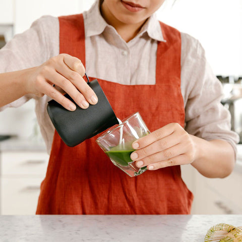 Person pouring green liquid from a black container into a glass, wearing a red apron.