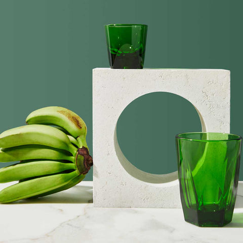 Green glass tumblers and bananas on a white surface with a green background