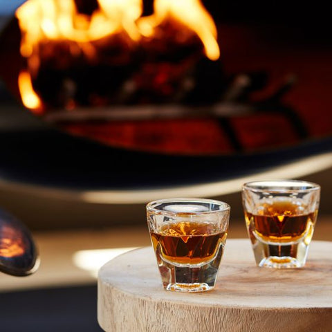 Two shot glasses with a dark liquid on a wooden table in front of a fireplace.