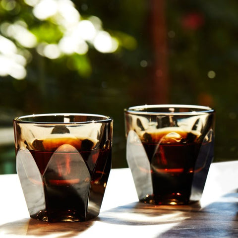 Two glasses of dark liquid on a table with a blurred natural background