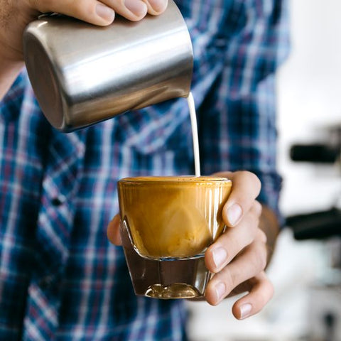 Person pouring milk from a metal pitcher into a glass cup held in the other hand.